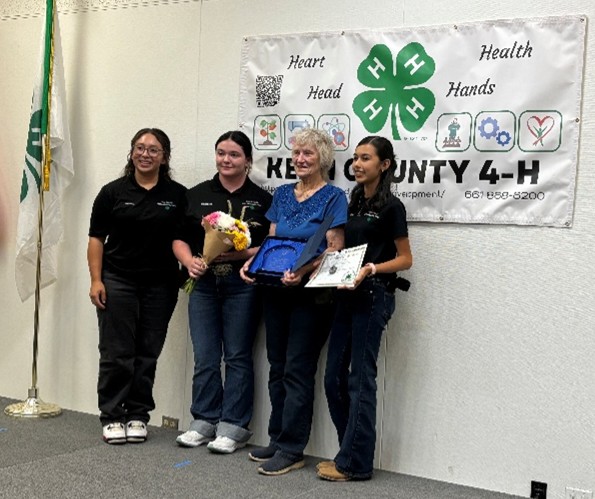 Kern County volunteer Jeanne Walford holding an award standing for photos with three other individuals, perhaps California 4-H youth members. One member is holding a bouquet of flowers, another is holding a certificate and all four are smiling standing in front of a Kern County 4-H banner.