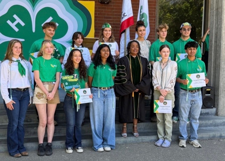 4-H high school graduates pose with Dr. Holmes at State Field Day. The youth are posing with their congratulatory certificates and honor cords, while Dr. Holmes is dressed in academic regalia. The group poses on steps in front of a 4-H Beyond Ready banner. 