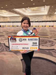 A 4-H youth member is shown smiling, holding a large 3rd place printed award representing a scholarship for Cub Bowhunter Female and a "The Vegas Shoot" medal, which is attached to a lanyard around their neck. The youth is indoors in a large conference room with a large circle pattern on the carpet.
