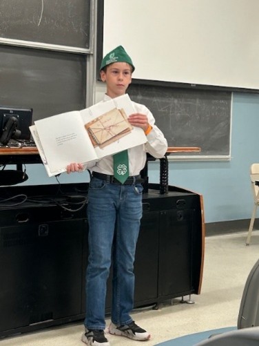 A 4-H youth member in 4-H attire standing at the front of a classroom, showing a book with various other papers. There are chalkboards and a desk in the background.