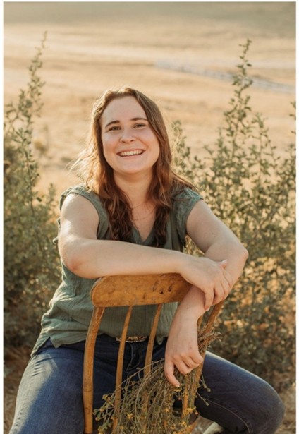 Photograph of Alexa D. sitting outdoors on a wooden chair turned backward, with arms resting on the chair's backrest. Background features dry grassland and green shrubs, highlighting a natural, rustic setting during daylight.