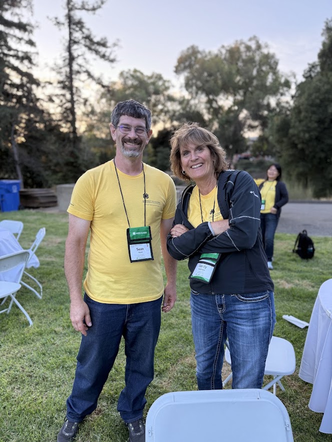 Two adult 4-H chaperones wearing yellow event shirts and name badges smile while standing together outdoors on a grassy area during an evening conference activity. White folding chairs and trees are visible in the background