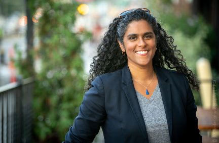 A woman with long dark curly hair smiles and poses wearing a blue fitted blazer