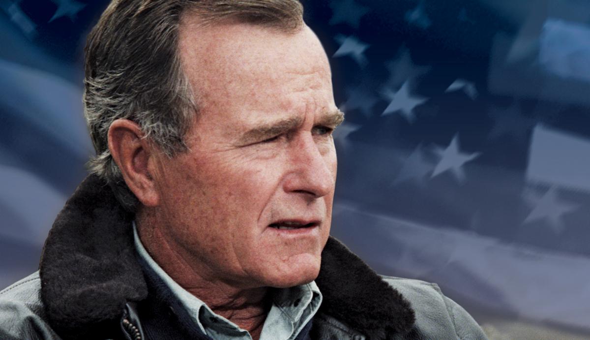 A man wearing a coat with a fur collar in the foreground with an American flag in the background