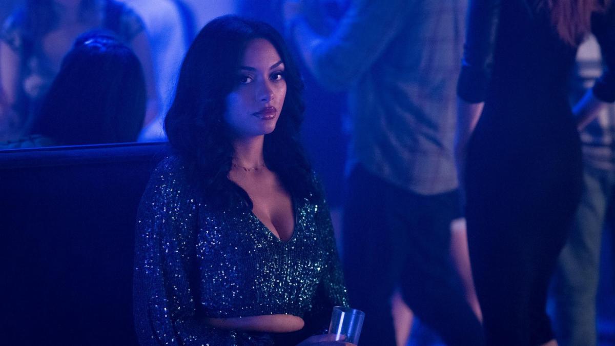 A woman with long dark hair and a sparkly dress sits in a club with people standing behind her