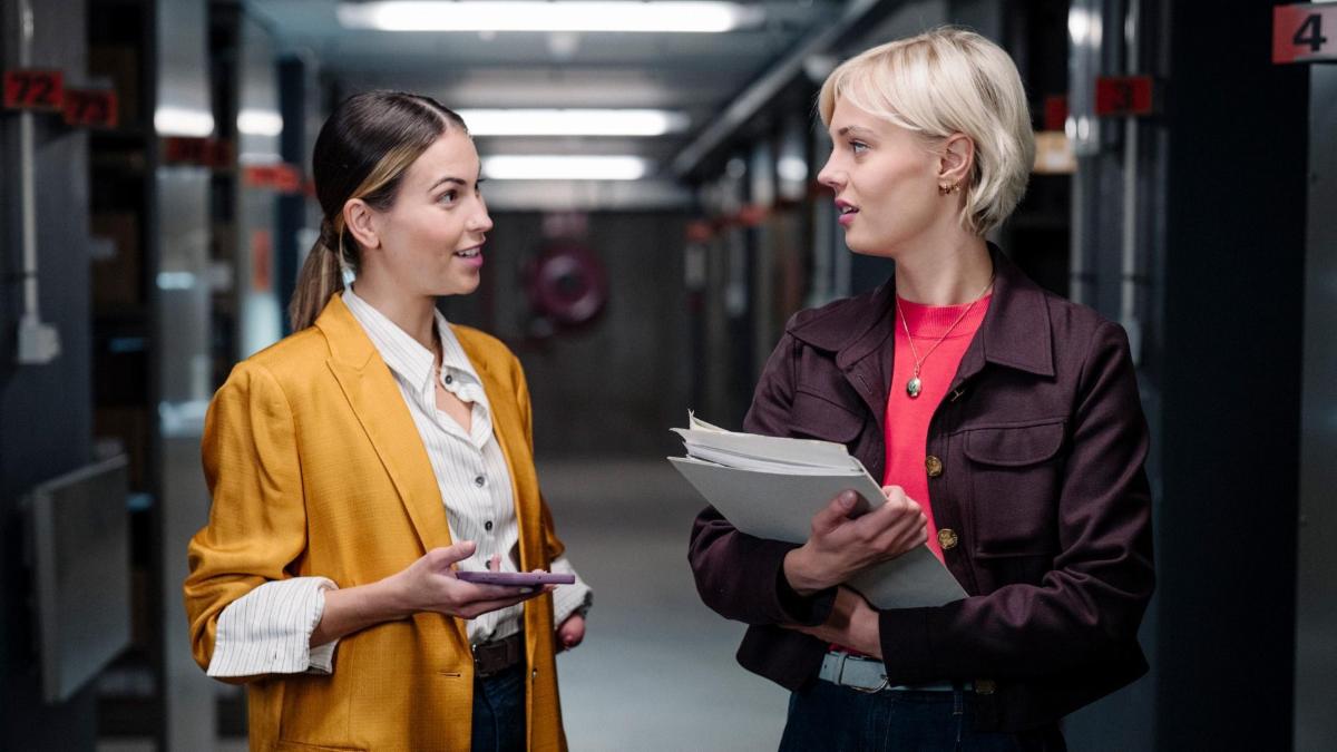 A woman with a yellow blazer talks to a woman in a burgundy wool coat in a hallway