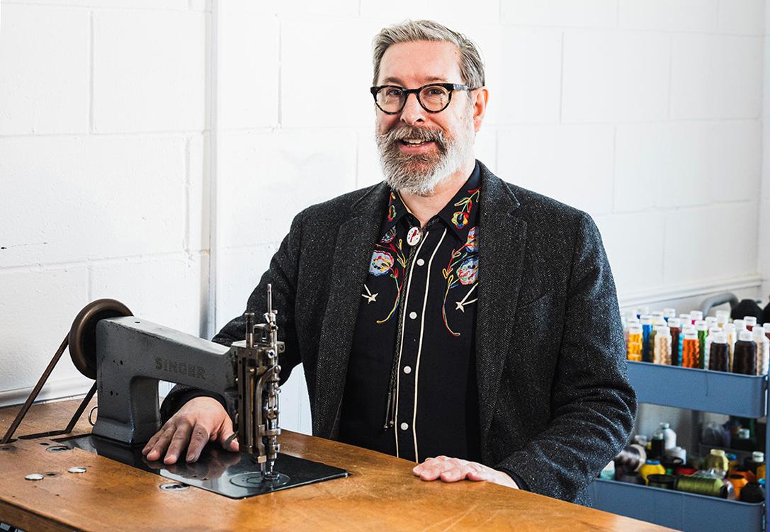 A bearded man wearing glasses and a black embroidered western shirt and blazer sits at a sewing machine