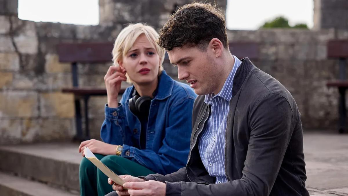 A man and woman sit in an outdoor stone structure, the man is reading a piece of paper