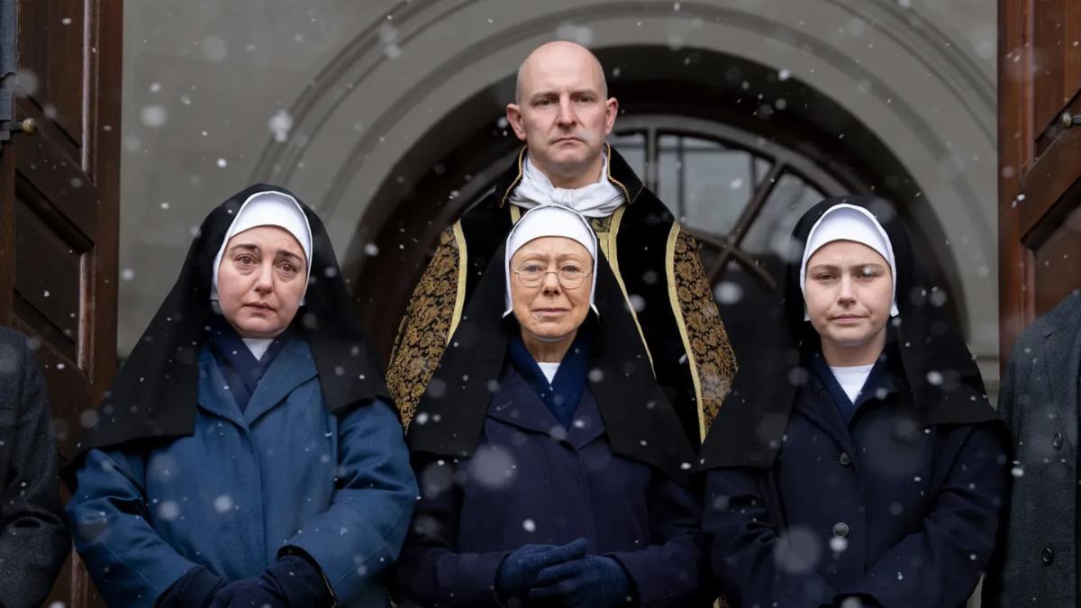 Three women dressed as nuns and man in a decorative coat look solemn while standing in a doorway with snowflakes
