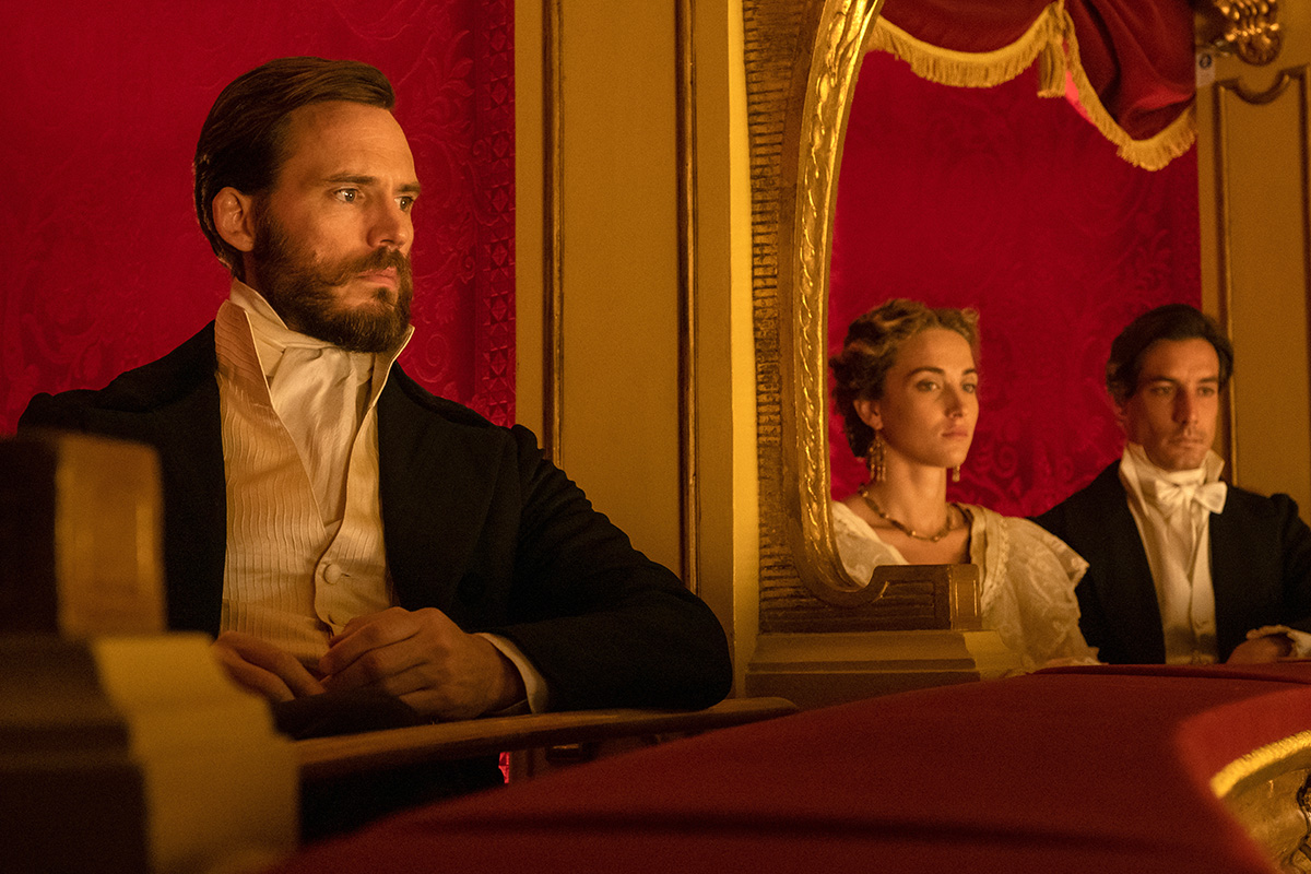 A bearded man in formal 19th-century attire sits in a richly decorated theater box with red velvet walls, while a woman and another man are visible in the next booth, all gazing toward the stage.