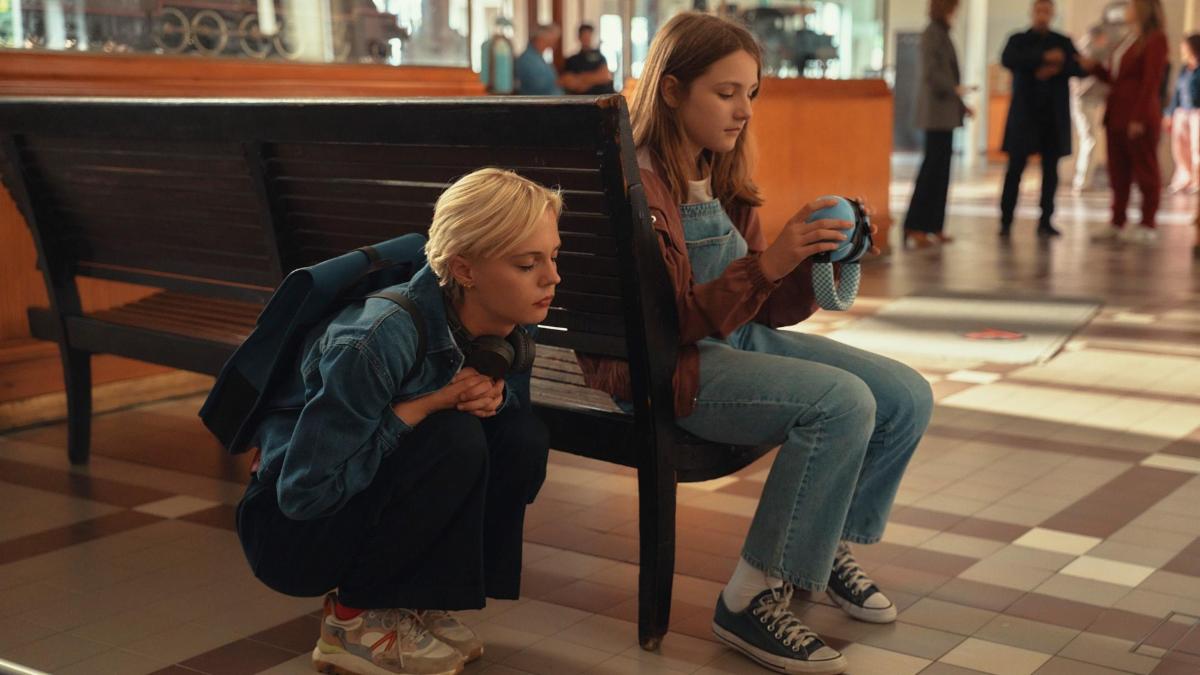 A woman crouches with her eyes closed behind a bench with a young woman holding a pair of headphones
