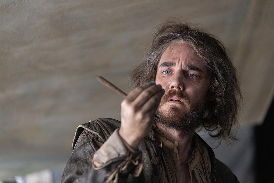 A man with disheveled hair and a dirty face looks down worriedly at something on his fingers
