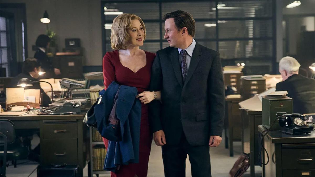A woman in a dark red dress links arms with a man in a suit in an office from the mid 1900s