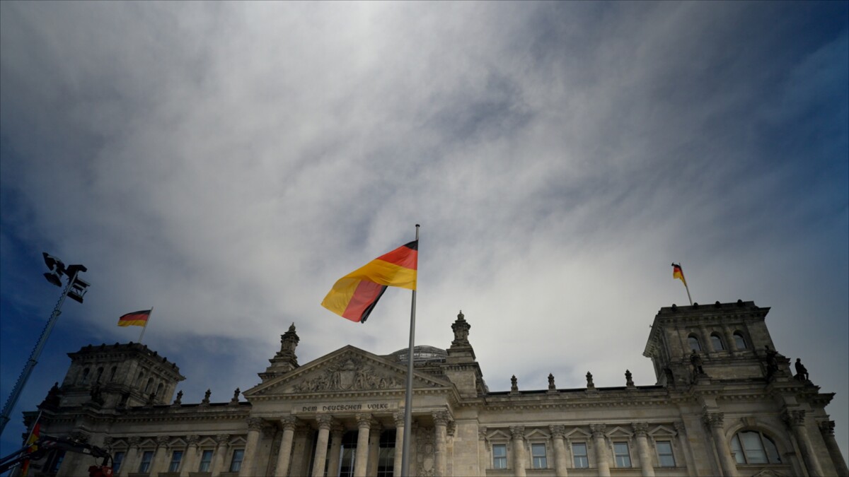 The German flag waves in front of the historic Reichstag building in Berlin under a cloudy sky.