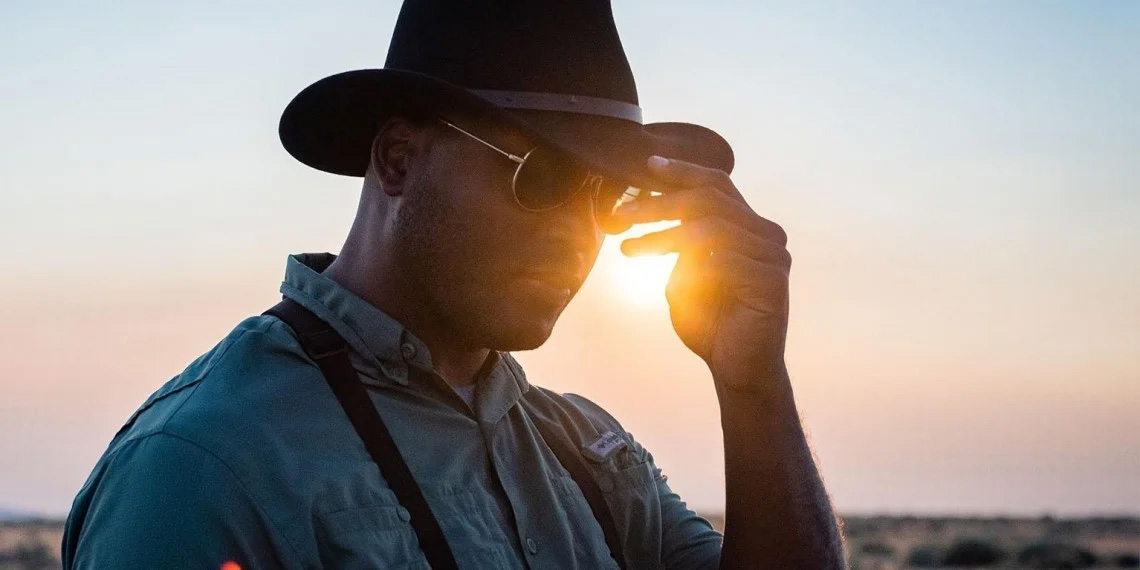 A man in a work shirt and sunglasses tugs on his black cowboy hat in almost silhouette in front of the sun