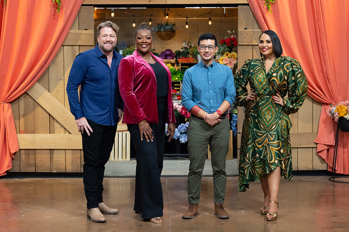 Two men and two women stand in front of a tv show set with wood doors, flowers and orange curtains