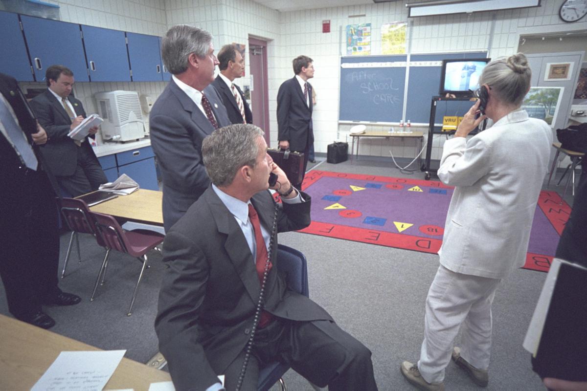 President Bush, men in suits and a woman on a phone in a classroom on a phone watching a TV with a tower on fire