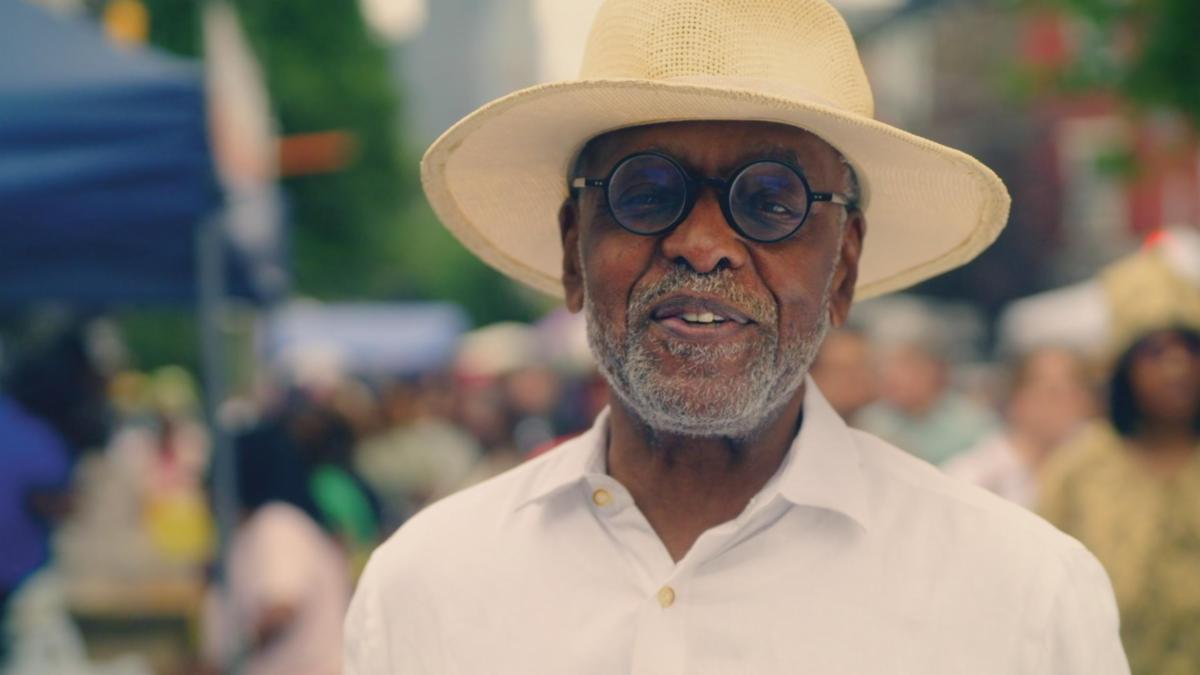 A man wearing a straw hat and glasses smiles with a busy sidewalk scene behind him