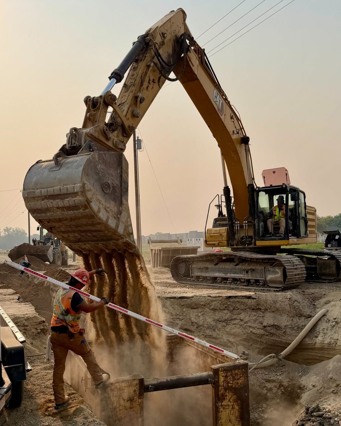 Excavator pours pipe bedding into a trench box while worker get ready to measure depth for Ustick Road project