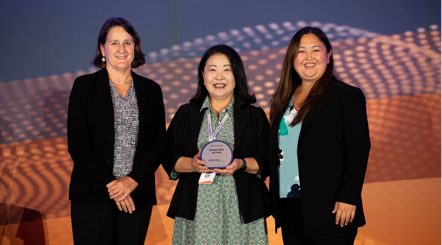 Center for Resource Solutions Chief Executive Officer Jennifer Martin stands to the left of Assistant General Managers Kelly Nguyen and Stacie Takeguchi in front of an orange backdrop. At the center, Nguyen holds an award.