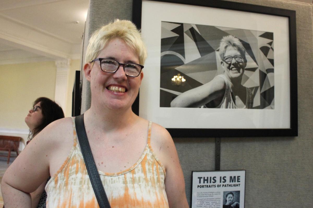 Leticia poses with her black and white portrait at the Portraits of Pathlight exhibit at the Boston State House. She has short blonde hair and glasses, wearing an orange dress.