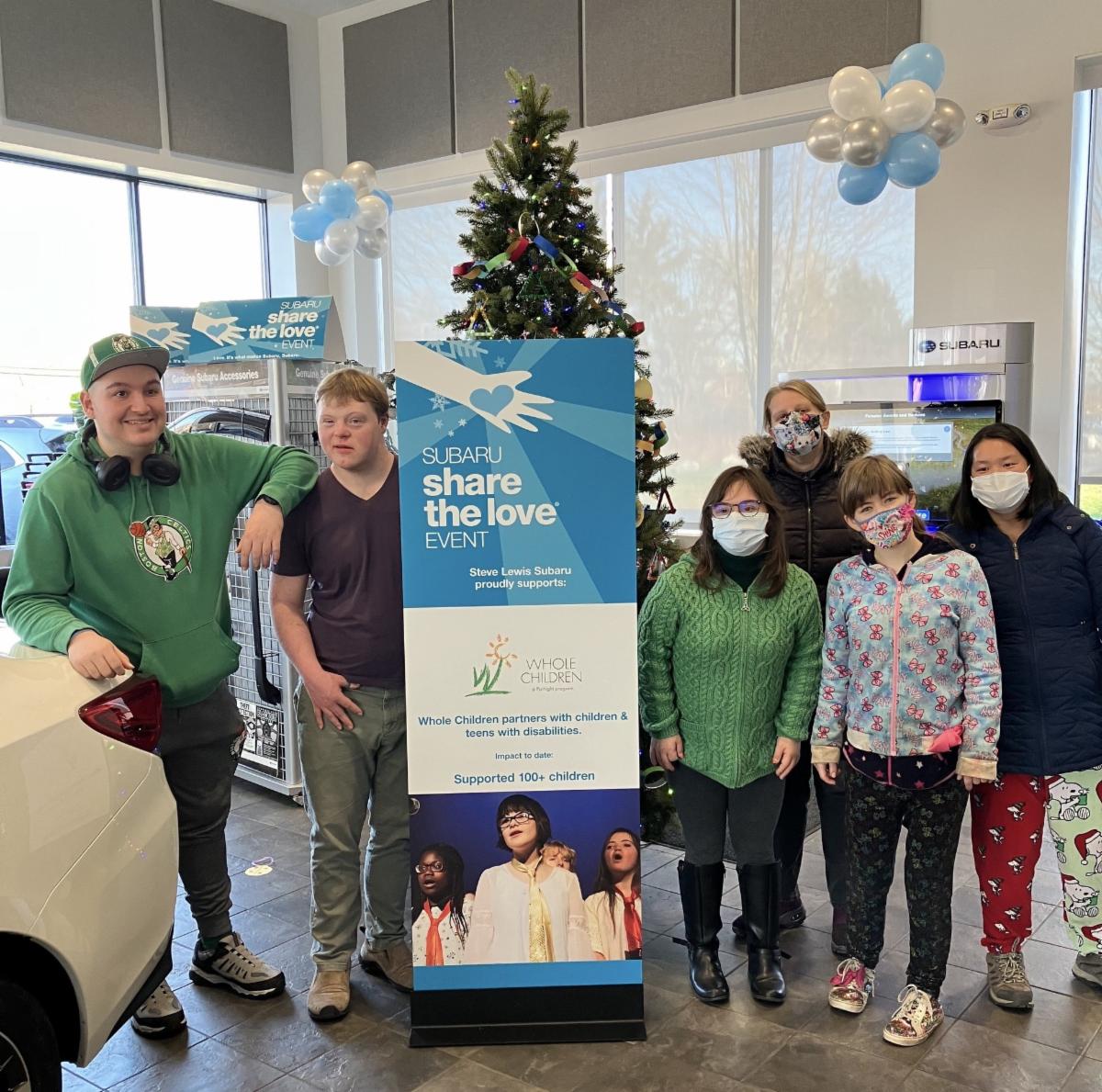 Several people stand in front of a sign for Subaru Share the Love campaign, with a holiday tree behind them. 