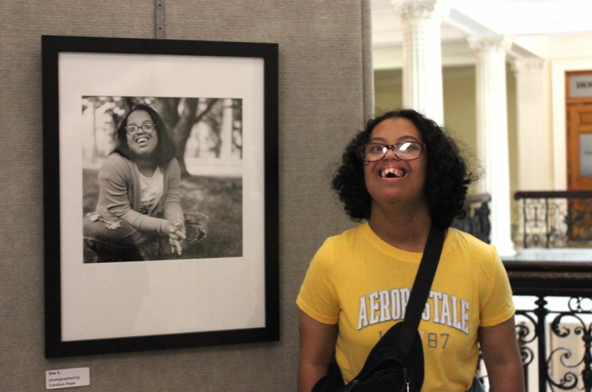 Zoe poses with her black and white portrait at the Portraits of Pathlight exhibit at the Boston State House. She has dark curly hair, glasses, and wearing a yellow t-shirt.