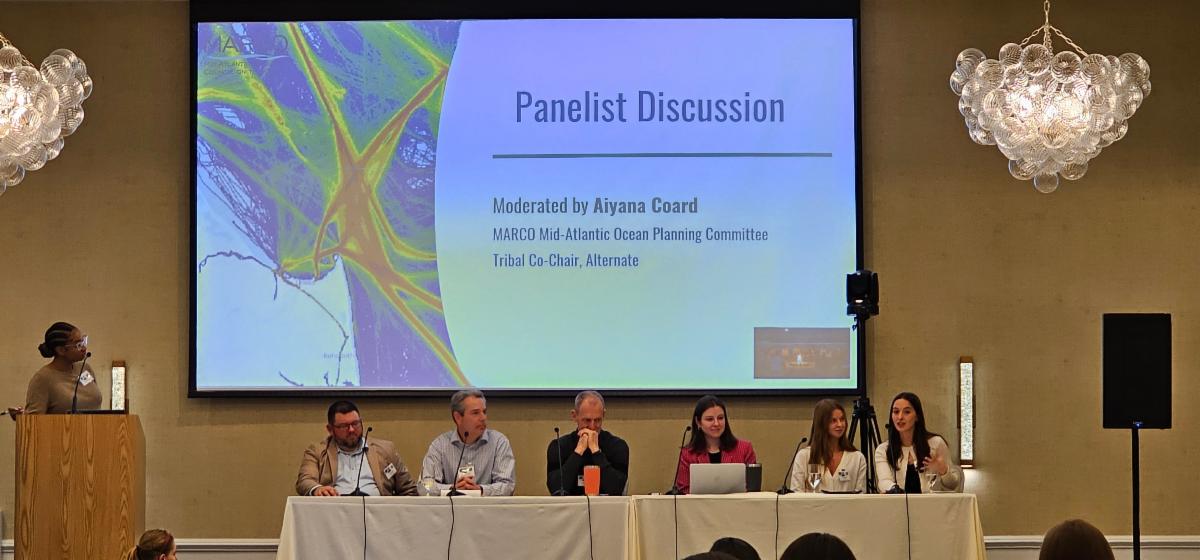 Six seated panelists with a slideshow on the wall behind them