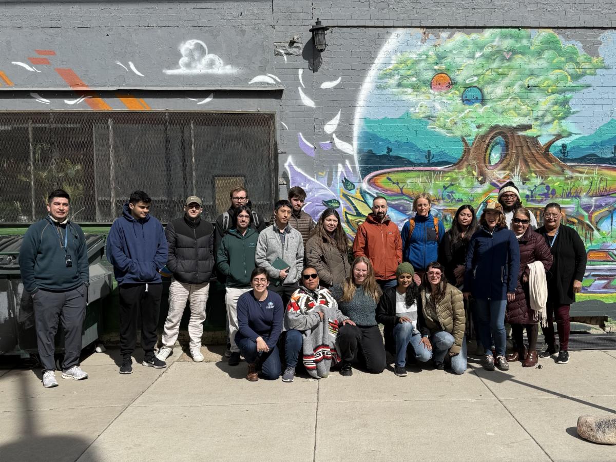A big group of people standing in front of a wall with a mural on it. 