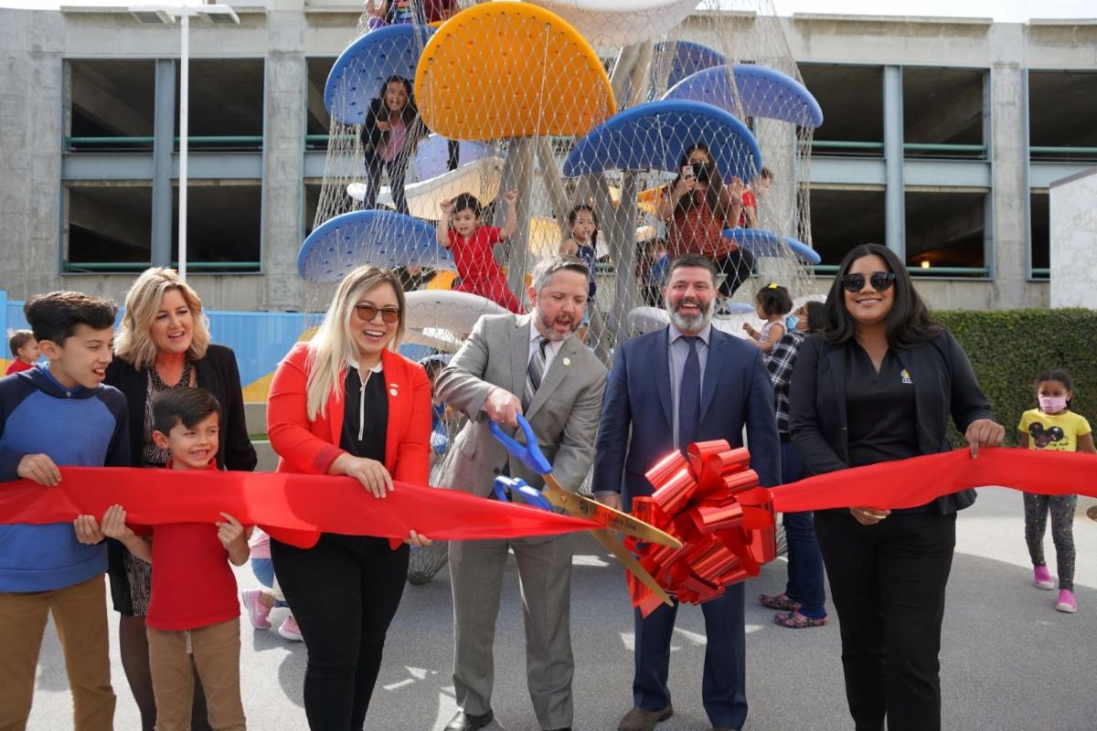 A picture with a group of people and children holding a red ribbon and a man with a big scissors cutting it. There are many children on the Luckey climber in the back.