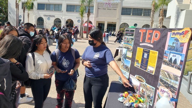 A picture of a group of people in front of a building. One of them is pointing at a big board that says TEP with many pictures on it.