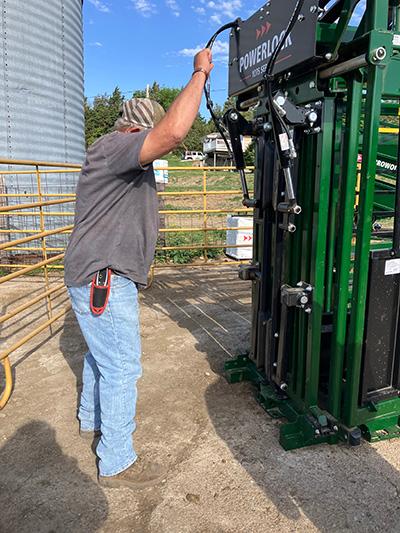A man in a grey shirt and cap stands outdoors reaching up to operate a lever on a large green piece of agricultural machinery labeled POWERLOCK with a tall silver grain bin visible in the background