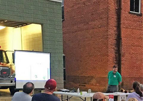 GA AgrAbility staff presenting a PowerPoint on a screen in Pike County outside of large gray building on left and large red-brick building on right