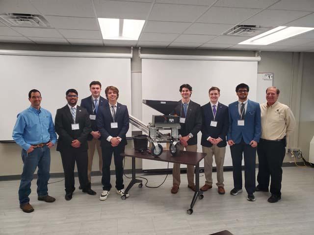 A group of young men in suits and two men in casual shirts stand in a classroom around a dark wooden table displaying a small mechanical wheeled prototype machine with a hopper on top