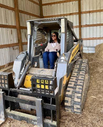 A woman wearing sunglasses and a patterned shirt smiles while sitting inside the operator cab of a yellow and black New Holland tracked skid steer loader parked inside a wooden barn