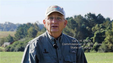 Field with large trees in background Gary Black commissioner of agriculture state of georgia wearing his white and grey baseball cap and a button down long sleeve shirt 