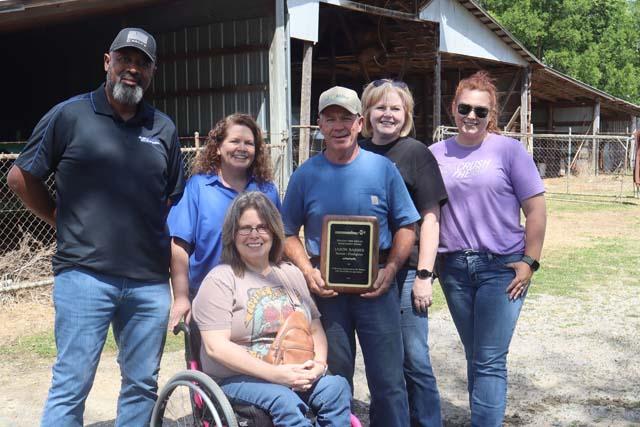 A group of six people stand outside a wooden barn smiling for a photo including a woman in a wheelchair in front and a man holding up a dark wooden award plaque