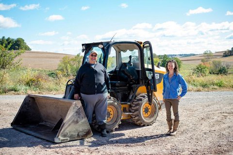 Garvin and Abbie in a beautiful quiet hilly area standing by a backhoe on gravel smiling facing the camera 