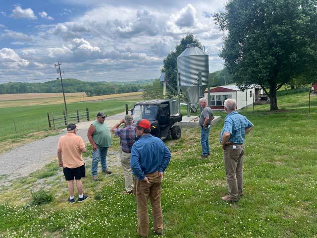 A group of men stand in a grassy area talking near a black utility task vehicle parked next to a tall silver metal feed grain bin with a farm house in the background