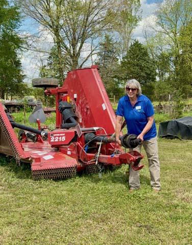 A smiling woman wearing sunglasses and a blue shirt stands in a grassy field next to a large red pull behind rotary cutter farm implement labeled 2215