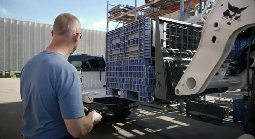 Man with back to camera using smartphone app to control skid-steer loading that is lifting a large plastic crate