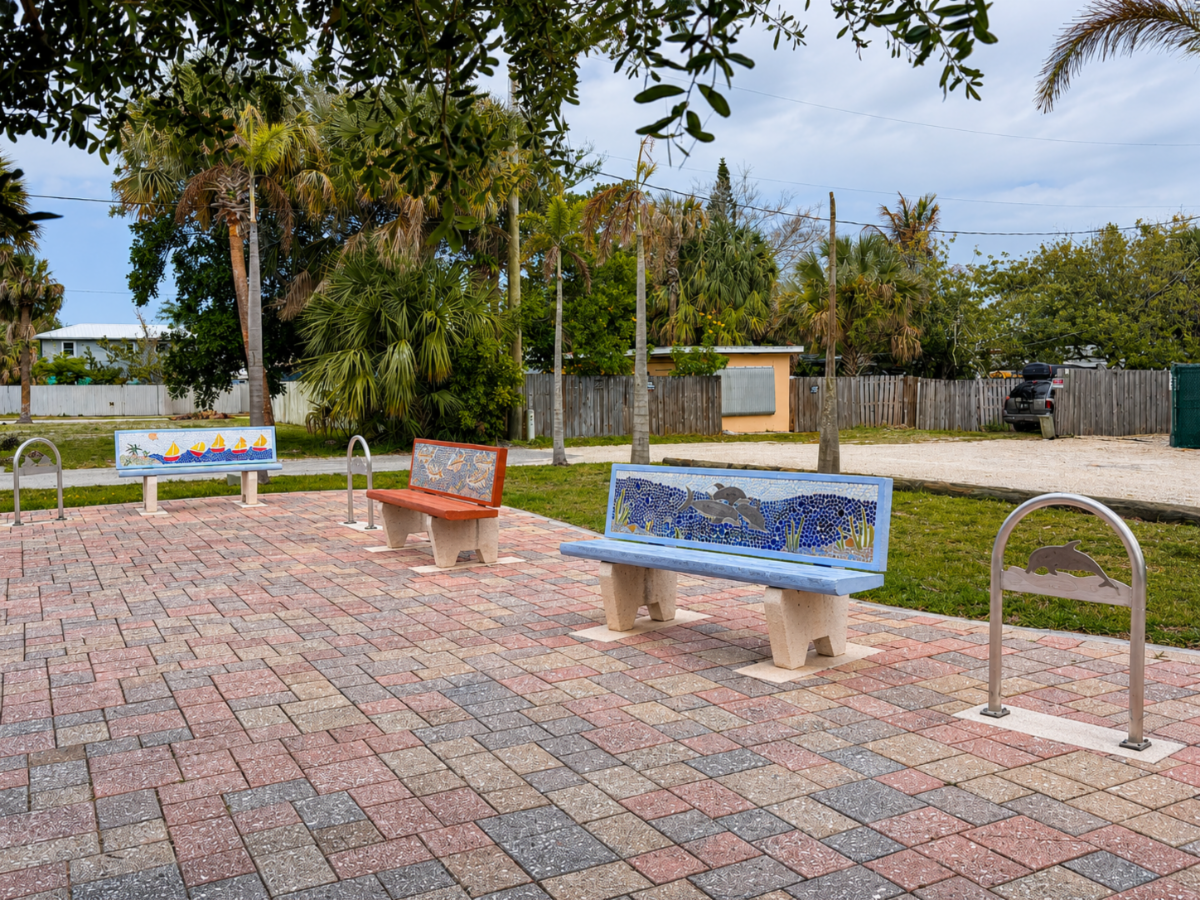 Three mosaic art benches and decorative metal bike racks installed on a brick-paved plaza in the Rio CRA, surrounded by palm trees and tropical landscaping.