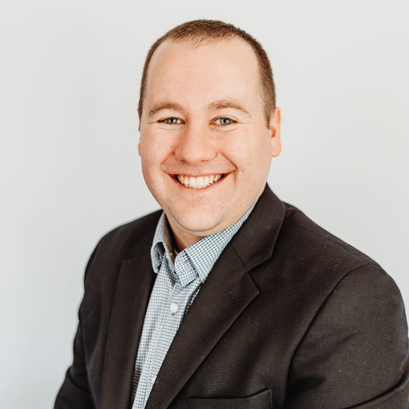 A mid-aged white man is smiling at the camera for a head-shot. He's wearing a dark suit jacket and light blue collared shirt.