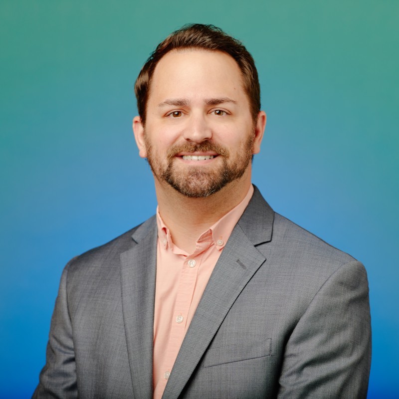 A headshot of Kasey Skala. He's a white man with facial hair and wearing a gray sports jacket and a coral collared shirt.