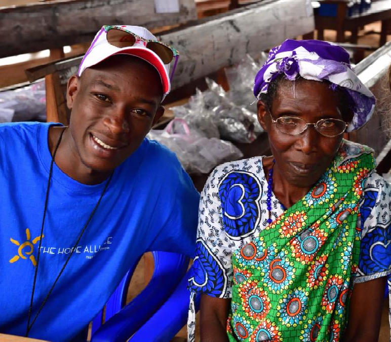 A Hope Alliance volunteer in a blue shirt and white cap sits beside an older woman wearing colorful patterned clothing and a purple-and-white headscarf. Both are seated closely together and looking toward the camera. The image captures a warm moment of connection and community engagement during an outreach or vision care program.