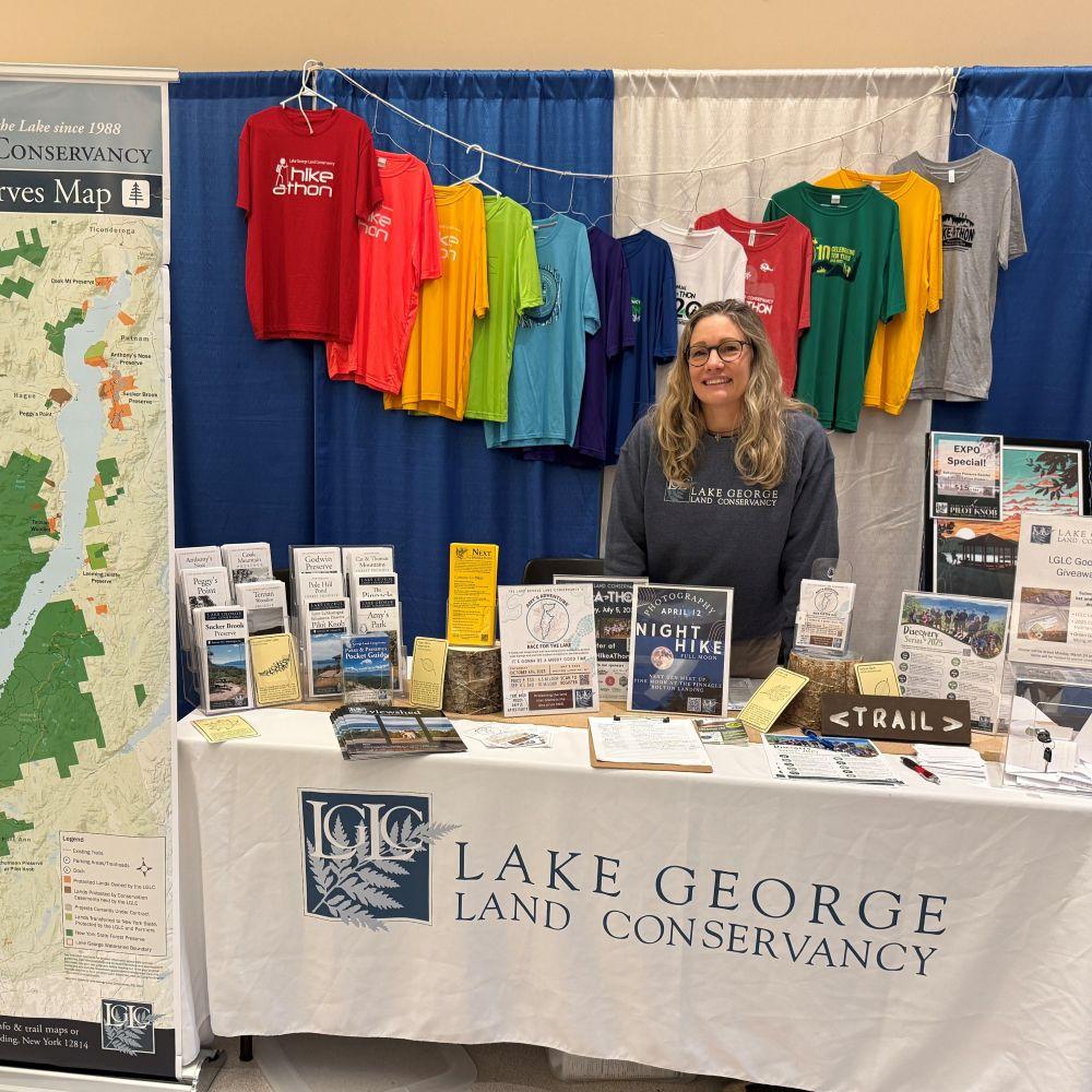 A woman stands behind a table with "Lake George Land Conservancy" printed on the tablecloth. Informational materials are displayed.
