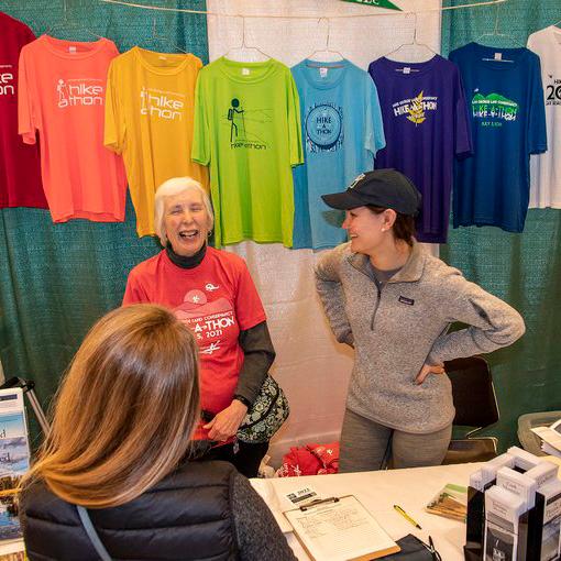 Two women laugh together. They are standing in front of colorful shirts, at a table with informational materials.