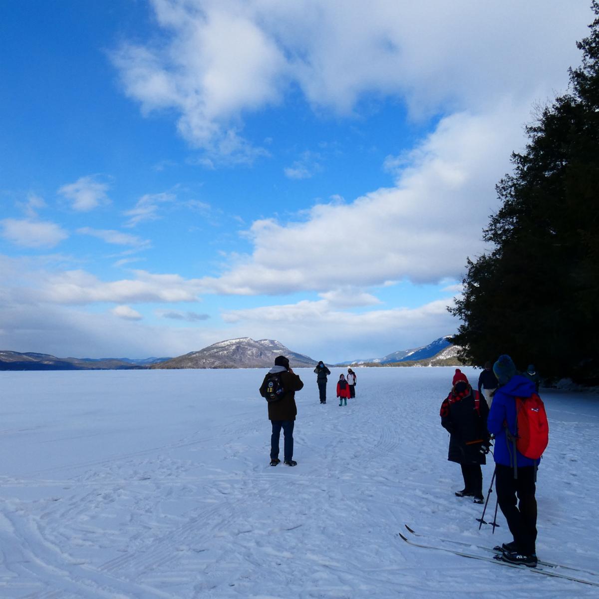 A few people stand on snow-covered iced-over lake, wearing snowshoes and skis. Snow covered mountains are in the distance.