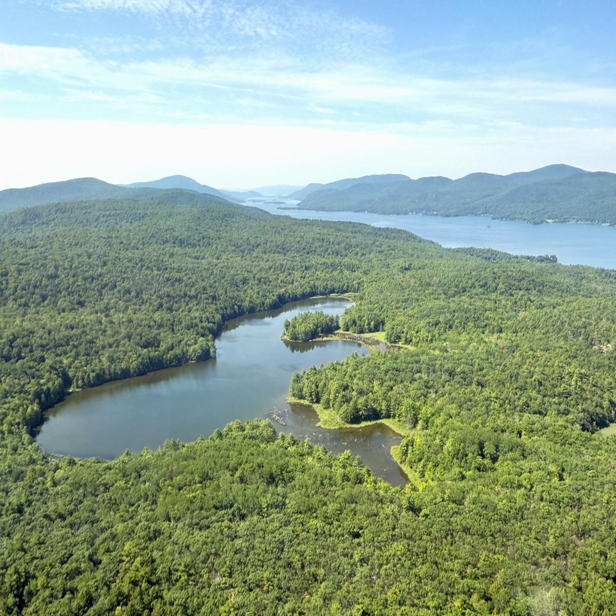 Aerial photo shows a pond surrounded by green forest and mountains, beside a lake.