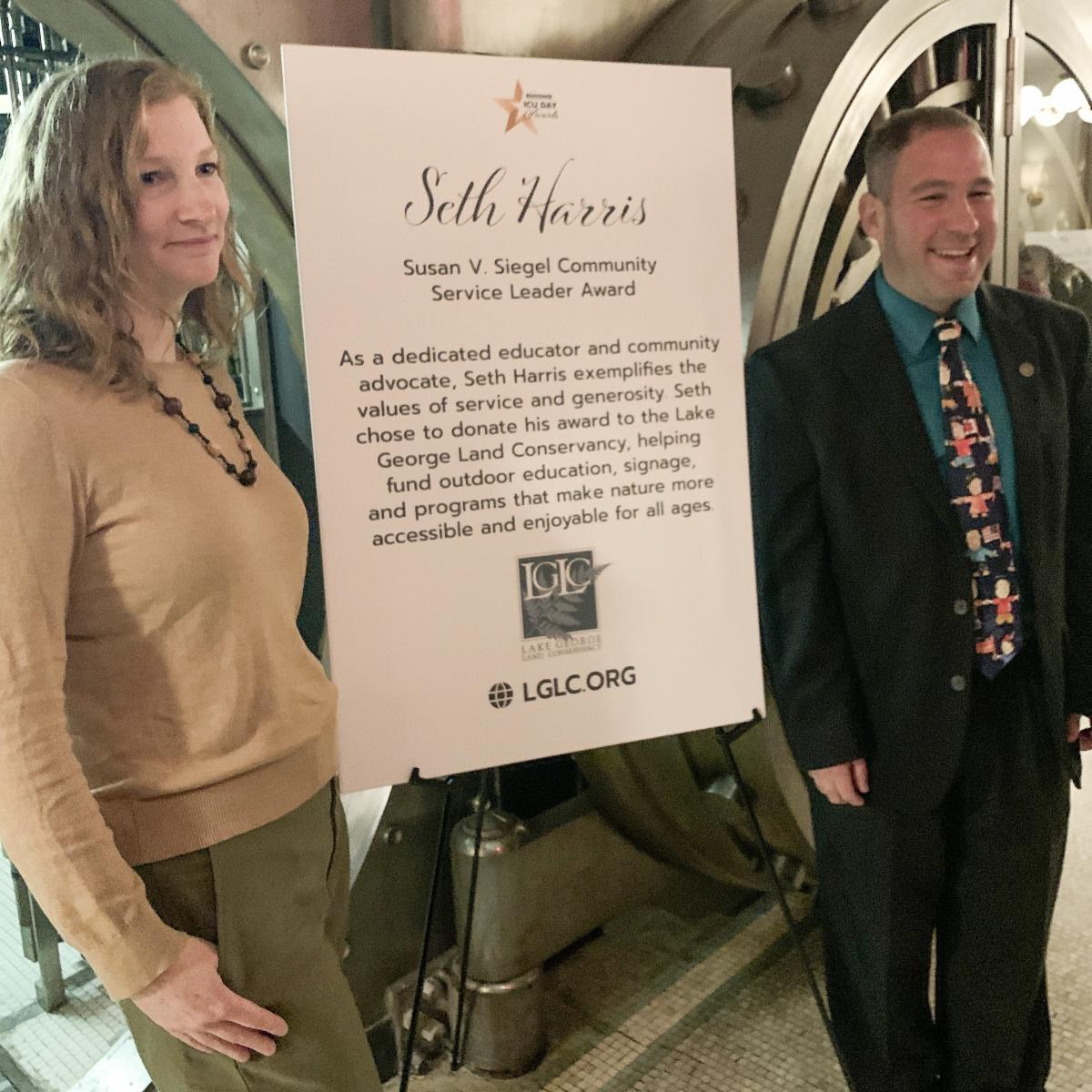 LGLC Communications Manager Sarah Hoffman and awardee Seth Harris stand next to a sign that display a description of the Susan V. Siegel Community Service Leader Award.
